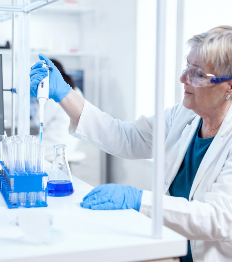 Senior scientist filling test tube wearing protective equipment using molecular dispenser. People in innovative pharmaceutical laboratory with modern medical equipment for genetics research.